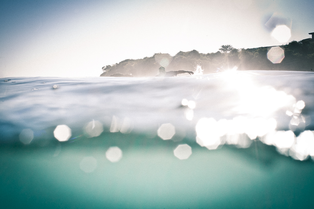A surfer paddles away from the camera, which is partially obscured by the water